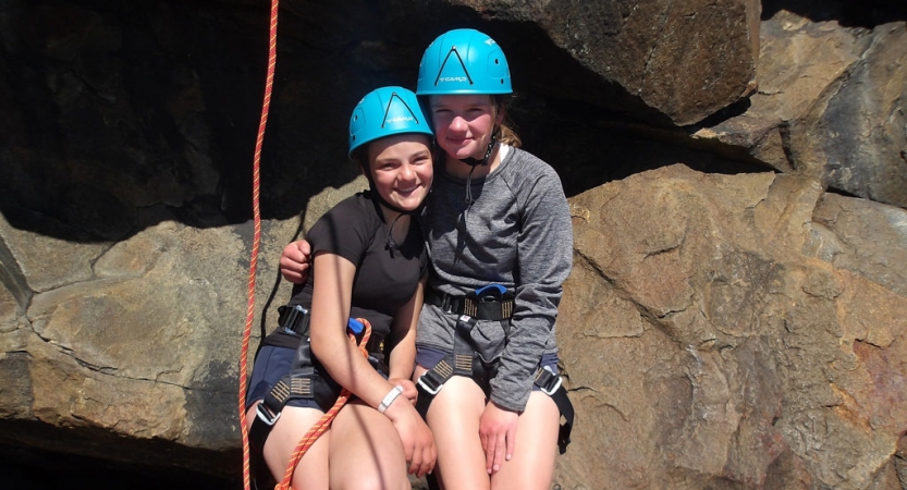 Two people wearing safety gear embrace at the base of a large rock. 